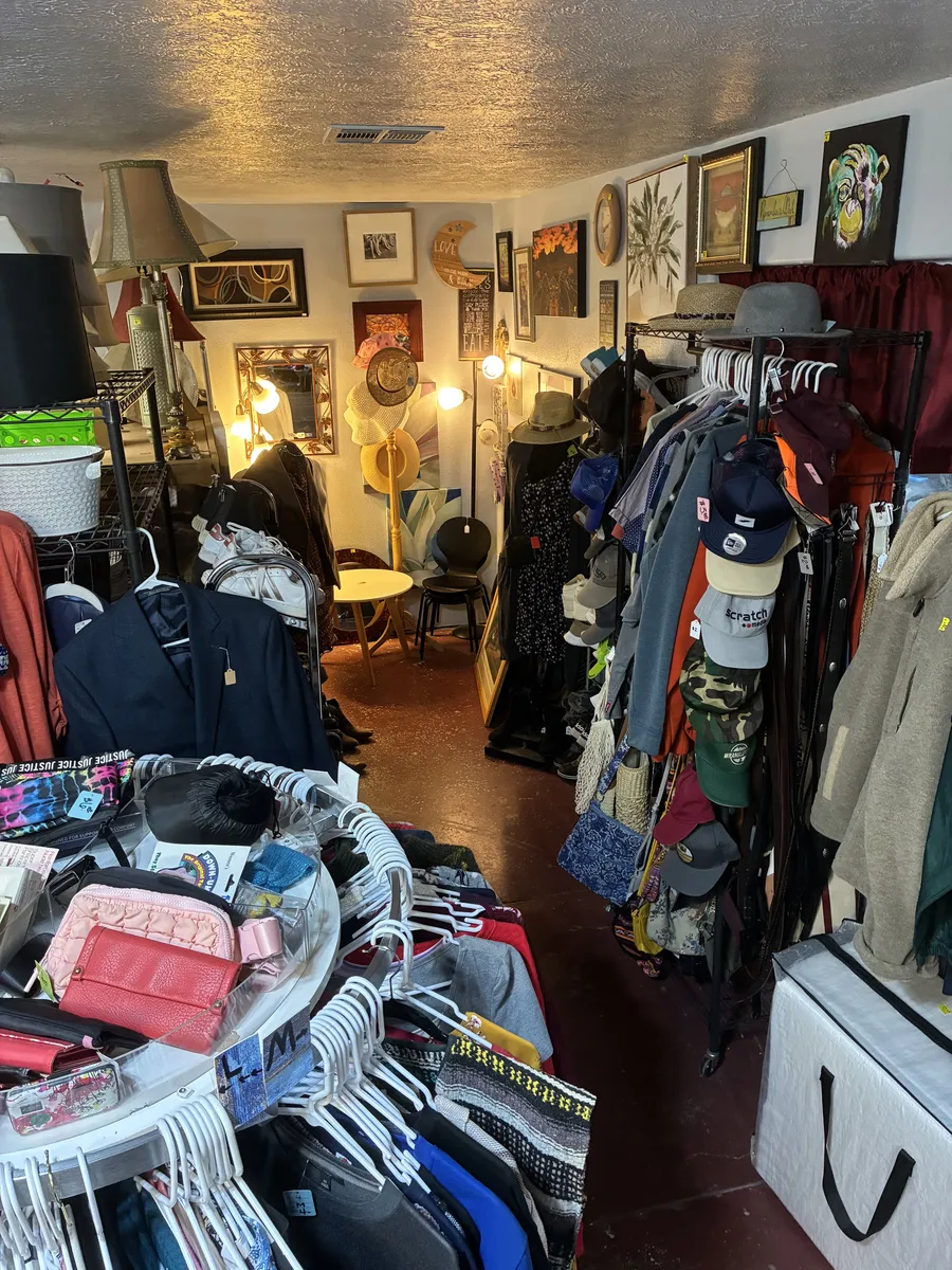 Clothing racks, bags and hats with framed art along the wall at BAM Thrift store