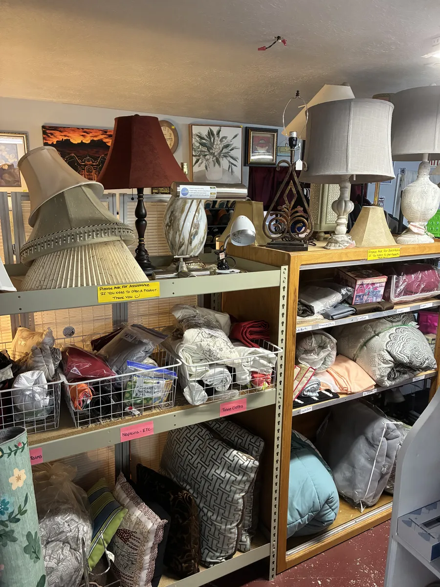 Lamps above shelves of pillows, yarn and folded textiles at BAM Thrift store